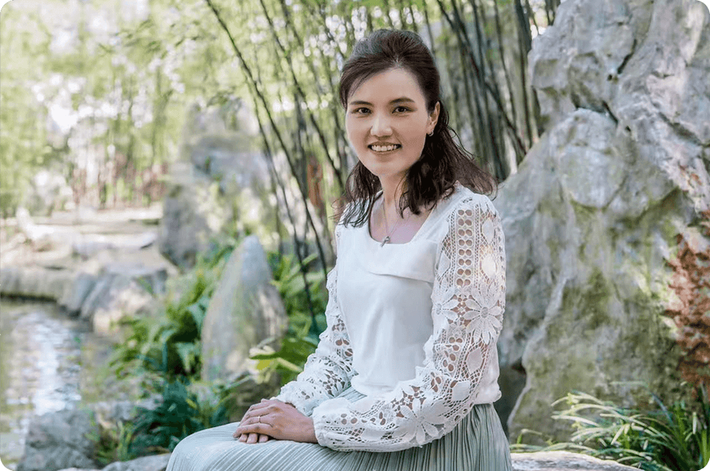 Smiling woman in white lace blouse sitting by rocks and bamboo in a serene garden setting with natural sunlight.
